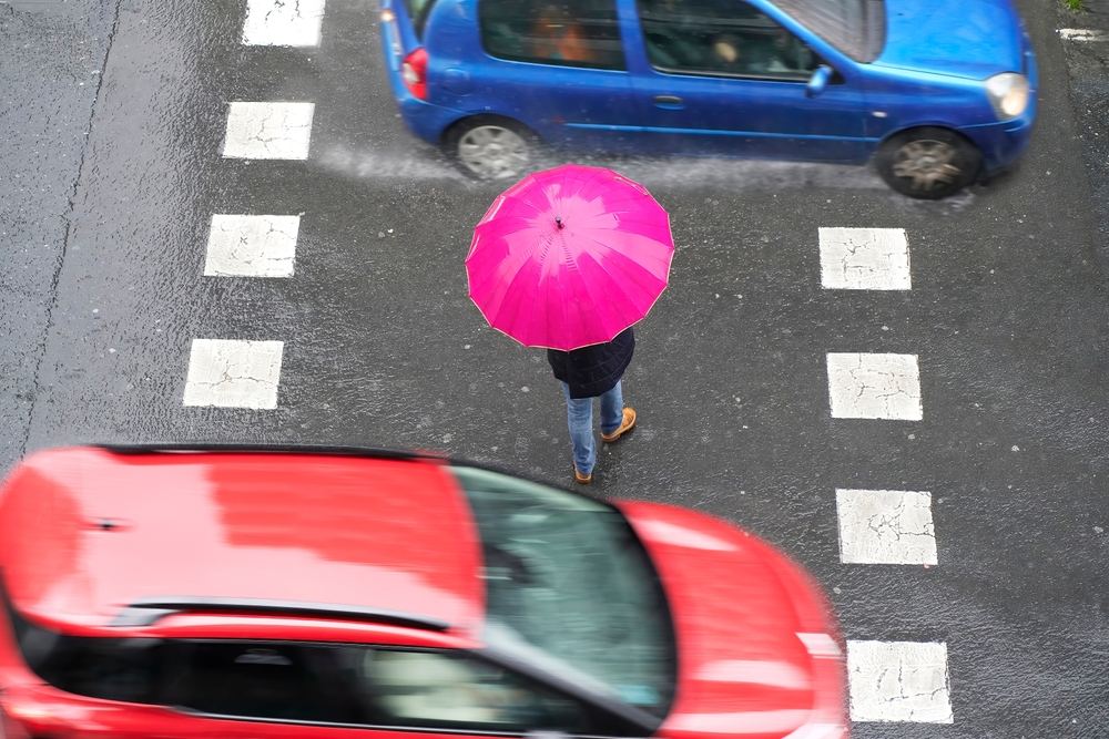 Aerial view of a pedestrian with a pink umbrella crossing a wet Wisconsin street between moving vehicles representing the danger and liability in a car hits pedestrian accident claim