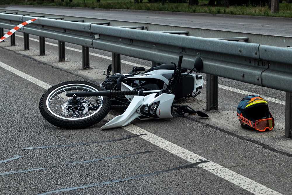 White motorcycle lying on its side by a highway guardrail after a crash, with a helmet and goggles resting on the pavement nearby